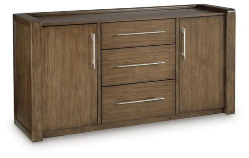 Three-quarter front view of Ashley Marxmore Dining Room Server (D863-60), featuring a rich brown wood-grain finish, three spacious center drawers, two side cabinet doors, sleek silver-tone handles, and contemporary buffet-style storage.