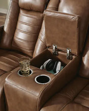 Ashley Gruaro Brown Power Reclining Loveseat (PCU1420018) with an open center storage console holding headphones, dual integrated cup holders, and generously cushioned reclining seats.