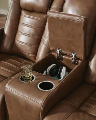 Ashley Gruaro Brown Power Reclining Loveseat (PCU1420018) with an open center storage console holding headphones, dual integrated cup holders, and generously cushioned reclining seats.