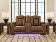 Ashley Gruaro Brown Power Reclining Loveseat (PCU1420018) displayed in an elegant living room with padded seating, center console, matching end tables, and blue LED base lighting.
