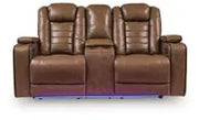 Straight-on front view of Ashley Gruaro Brown Power Reclining Loveseat (PCU1420018), showcasing dual cushioned seats, padded armrests, center console with cup holders, and integrated blue LED base lighting.