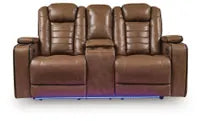 Straight-on front view of Ashley Gruaro Brown Power Reclining Loveseat (PCU1420018), showcasing dual cushioned seats, padded armrests, center console with cup holders, and integrated blue LED base lighting.