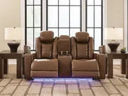 Ashley Gruaro Power Reclining Loveseat (PCU1420018) in a contemporary living room, featuring two plush reclining seats, a center storage console, cup holders, and blue LED floor lighting.
