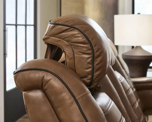Side-angle close-up of the Ashley Gruaro Brown Power Reclining Loveseat (PCU1420018), emphasizing its thick cushioned headrests, sculpted upper back supports, brown upholstery, and decorative black piping.